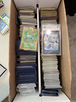 Two cardboard card storage boxes filled with stacks of standard-size Pokémon cards. Left box holds cards in colored penny sleeves and a few in top loaders, including a graded slab card and a first edition Base Set Venusaur. Right box contains a large stack of unsleeved cards