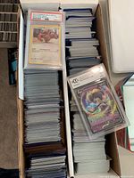 Photo shows two long boxes filled with sleeved Pokémon cards. Two graded cards are displayed on top of card stacks: a PSA graded Lickitung card and a BGS graded Pokémon V card.