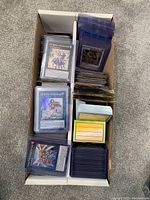 Box containing multiple stacks of Yu-Gi-Oh and Digimon trading cards, many in plastic sleeves and organized in smaller stacks or packs.