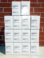 Photograph of 14 stacked white boxes labeled CF280A(II) (HP 80A) by NuPrint, each box containing a black toner cartridge, arranged in a pyramid shape against a brick wall.