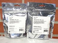 Two sealed silver-colored foil packages containing Canon PG-210 black ink cartridges placed side by side on a brick surface.