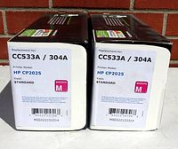 Photo shows two original boxed HP printer toner cartridges CC533A / 304A in magenta for HP CP2025 printer models with standard page yield.