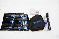 Photo showing six black individually packaged pre-moistened lens cloth wipes, a small spray bottle, a soft cloth, a cleaning brush, and a small black Zeiss branded pouch on white background.
