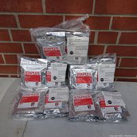 Lot of multiple silver sealed packages of Canon PGI-5 black ink cartridges with Staples branding, stacked in front of a brick wall.