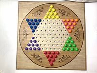 Top view of square wooden Chinese Checkers board with multicolored pegs arranged in starting positions. Board has holes and floral decorations.