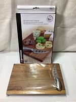 Mini acacia wood cutting board shrink-wrapped with stainless steel cheese knife on top and retail packaging box behind