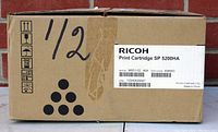 Brown cardboard box labeled Ricoh Print Cartridge SP 5200HA, model M851-03 with barcode and serial number. The box includes handling symbols and handwritten '1/2'.