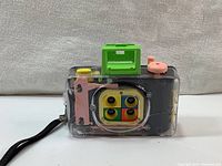Front angle showing the transparent case, four colorful lenses, pink shutter release, and green viewfinder of the camera.