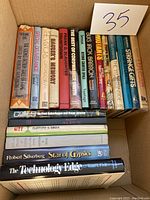 Photo of the collection of 19 hardcover books stacked in two rows inside a cardboard box with label number 35, spines visible showing various titles and authors.