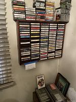 Photo showing a wooden hanging cassette rack on wall filled with multiple audio cassette tapes of mixed genres.