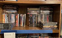 Wooden bookcase shelf with approximately 80 DVD movie cases arranged in several rows and stacks. Visible titles include various film genres and recognizable movie titles.