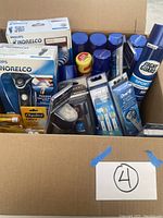 Box with 2 Philips Norelco electric shavers in original packaging, several Oral-B toothbrush replacement heads, multiple ChapStick lip balms, multiple Right Guard Sport deodorant cans, and additional grooming products.