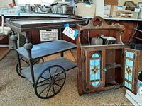 Photo of two vintage furniture pieces: a stained glass wooden wall cabinet with two doors opened displaying shelves inside, and a vintage rolling serving cart with three shelves and large spoked metal wheels.