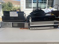 Front view of Pioneer LD-700 LaserDisc Player, Panasonic Magnavox player, and Akai video component with AV cables on top, shown on a white table outdoors.
