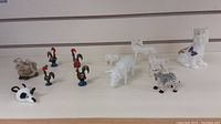 Photo of twelve small animal figurines arranged on a white surface, including roosters, pig, cat, horse, cow head, and zebra.