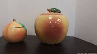 Two apple-shaped cookie jars, larger one approximately 9 inches tall and smaller one approximately 6 inches, displayed with lids on and off to show each jar and lid details.