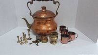 Full shot of copper kettle, four copper cups, five brass candlesticks, vintage brass padlock with keys arranged on white backdrop