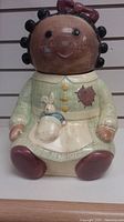 Front view of the ceramic cookie jar shaped like a seated girl with dark skin, black curly hair, green dress, red shoes, and a bunny in pocket.