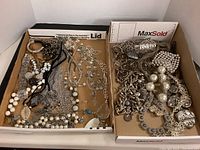 Two trays of silver colored costume jewelry with necklaces, bracelets, earrings arranged inside the boxes.