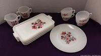 Group showing four rectangular plates stacked, cups, creamer, sugar bowl and one round plate all with red and orange floral designs on white glass.