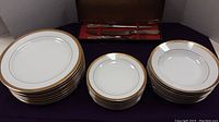Full view of stacked gold rimmed dinner plates, side plates and bowls arranged with carving set in background