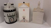 Three architectural ceramic cookie jars shaped as a castle, house and church, displayed side by side against a white background.