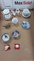 View of miniature bowls, cups, saucers, small vase, trinket container, decorative watering can, and cat figurines arranged in cardboard box with MaxSold branded backdrop.