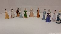 Wide angle photo showing all 12 miniature lady figurines in a row, showcasing variety of colors, dresses, and styles.