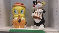 Front view showing both cookie jars side by side with Tweety on left wearing red hat and holding sunflower, Sylvester on right holding birdcage.