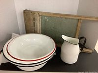 Three large enamel bowls stacked, enamel water pitcher, and vintage washboard with wooden frame and metal scrubbing surface