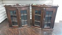 Pair of matching wood hanging display cases with glass paneled doors and carved decorative wood crest at top.