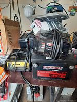 Multiple battery chargers stacked with cables wrapped around, including Motomaster and Noco Genius G7200 commercial charger.