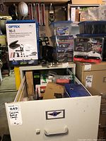 Wide view showing boxed Optex accessory kit, Solar Shades kit, storage containers, and Peak backup camera box on a shelf among other items.