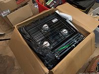 Photo showing the black 3-burner Atwood Wedgewood gas stove partially wrapped in black plastic inside an open cardboard box with foam protectors on the sides.