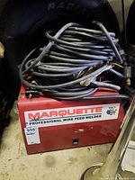 Front view of red Marquette professional wire feed welder with cable coiled on top, showing brand label and condition tag.