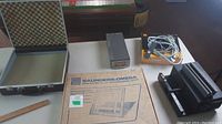 Photo showing black slide projector, carousel slide tray, rectangular electronic unit with cables, black case with foam interior, and box for enlarging easel.
