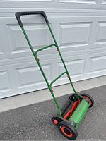 Full view of the manual lawn reel mower showing green frame, red reel blades, black rubber wheels with red centers, and padded black handle grip.