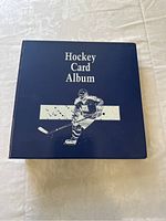 Closed front cover of blue Hockey Card Album binder with an image of a skater and text, placed on a patterned tablecloth.