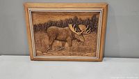 Framed wood carving art showing a moose in a marsh landscape with a background of trees.