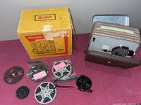 Kodak Brownie 8mm projector with original box and multiple 8mm film reels on pink fabric surface.