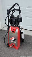 Full view of CANAD-X pressure washer with hose reel and spray gun, showing device standing upright next to a white garage door.