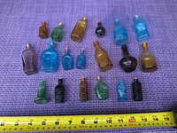 Photo showing 18 miniature bottles of various colors and shapes laid out on a purple textured surface with a tape measure showing length from 0 to approx 17 cm for scale.
