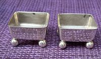 Two small square sterling silver condiment bowls displayed on purple woven fabric background.
