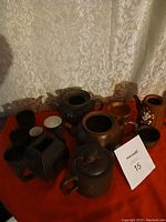 Photo showing six teapots and several small cups arranged on a red surface against a curtain background, displaying different shapes, colors, and designs.