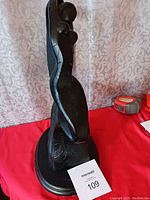 Dark brass abstract sculpture of a lady on an oval base, placed on a red cloth with a MaxSold auction tag in front.