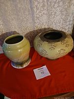 Photo showing two ceramic vases on red cloth, the green glazed heavier vase on the left, the beige floral raised pattern vase on the right