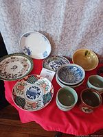 Photo showing collection of various ceramic plates and cups with different decorative patterns and colors arranged on a red cloth.