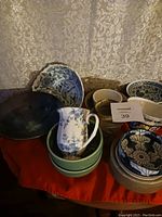 Various ceramic bowls, plates, and a small pitcher with blue and white Chinese floral designs along with plain colored ceramic bowls stacked together on a red cloth.