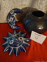 Photo showing blue ceramic crescent moon and sun decorations with white floral face details, and the round blue ceramic vase on a reddish surface with a MaxSold lot card.