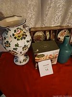 First photo showing large floral ceramic vase, set of small plates in wooden box, turquoise vase, and boxed item.